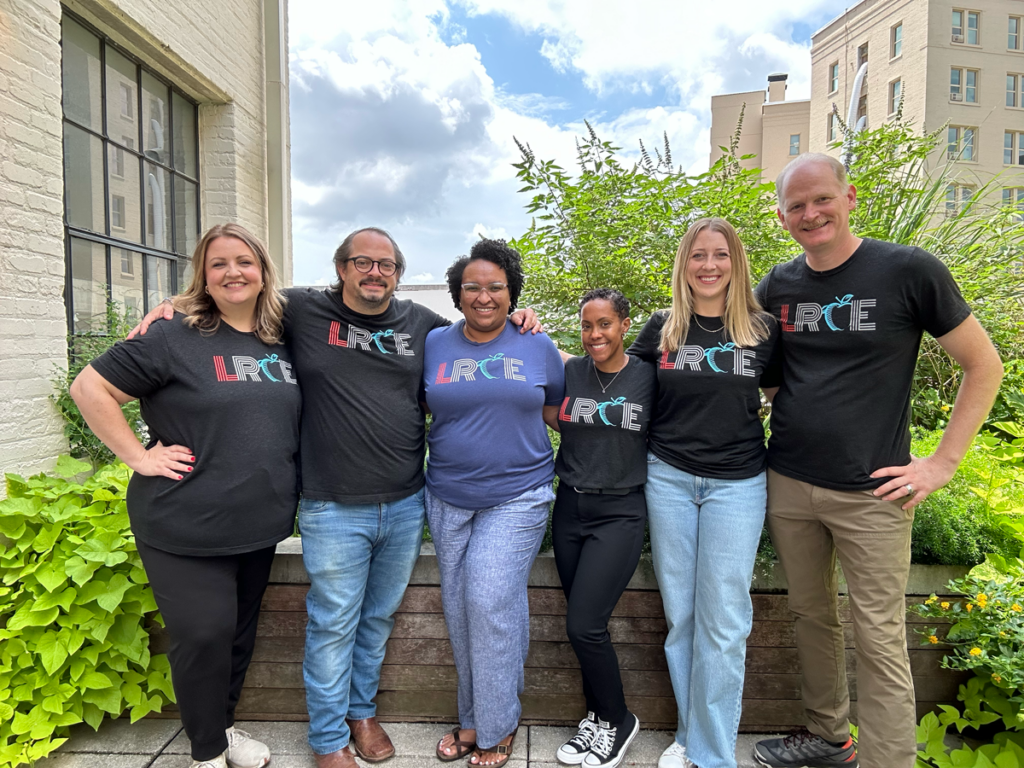 A group of six smiling LRCE team members stand together outside in front of greenery and city buildings, wearing matching LRCE logo t-shirts. Their camaraderie reflects the supportive community behind Louisiana Resource Center for Educators.