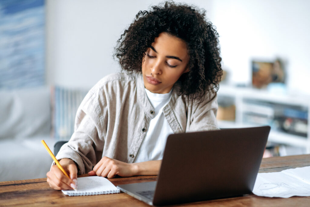 Woman studying remotely taking LRCE program while sitting in living room at laptop and taking notes