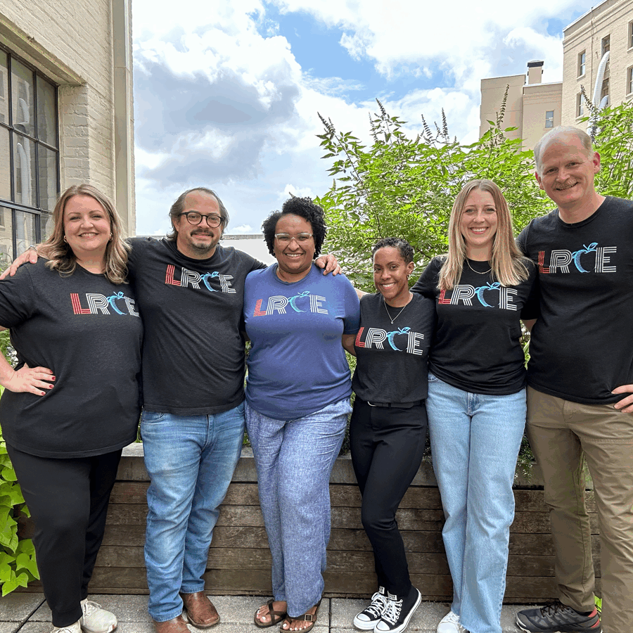 A group of six smiling LRCE team members stand together outside in front of greenery and city buildings, wearing matching LRCE logo t-shirts. Their camaraderie reflects the supportive community behind Louisiana Resource Center for Educators.