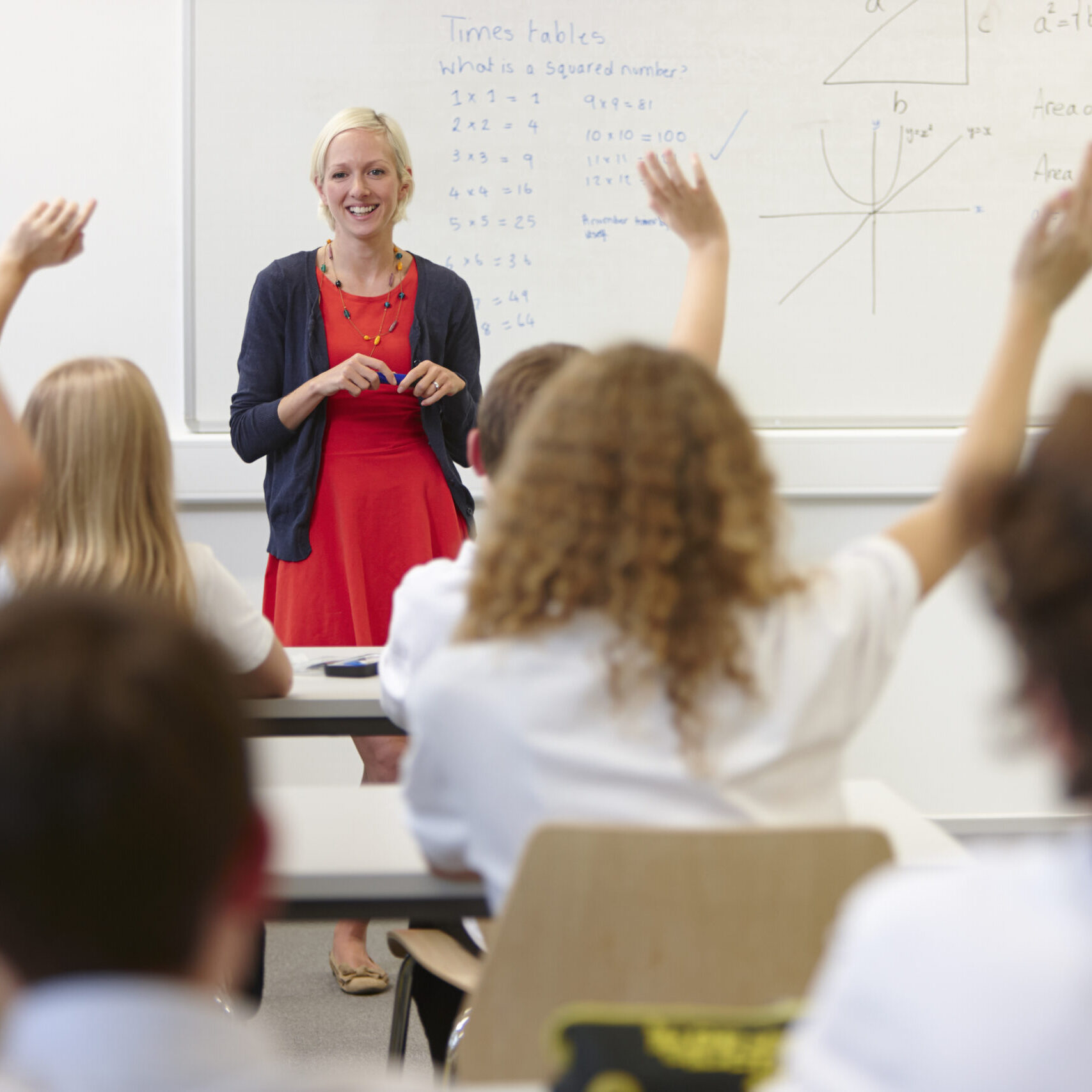 LRCE-Teacher-in-Classroom-with-Students A teacher smiles at the front of a classroom as several students eagerly raise their hands to answer a math question. The interactive and welcoming environment reflects the impact of LRCE-trained teachers.