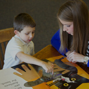 An LRCE-certified teacher in a blue sweater sits with a young student as they read a colorful picture book together. The child points to a page while the teacher listens attentively, encouraging early literacy skills.