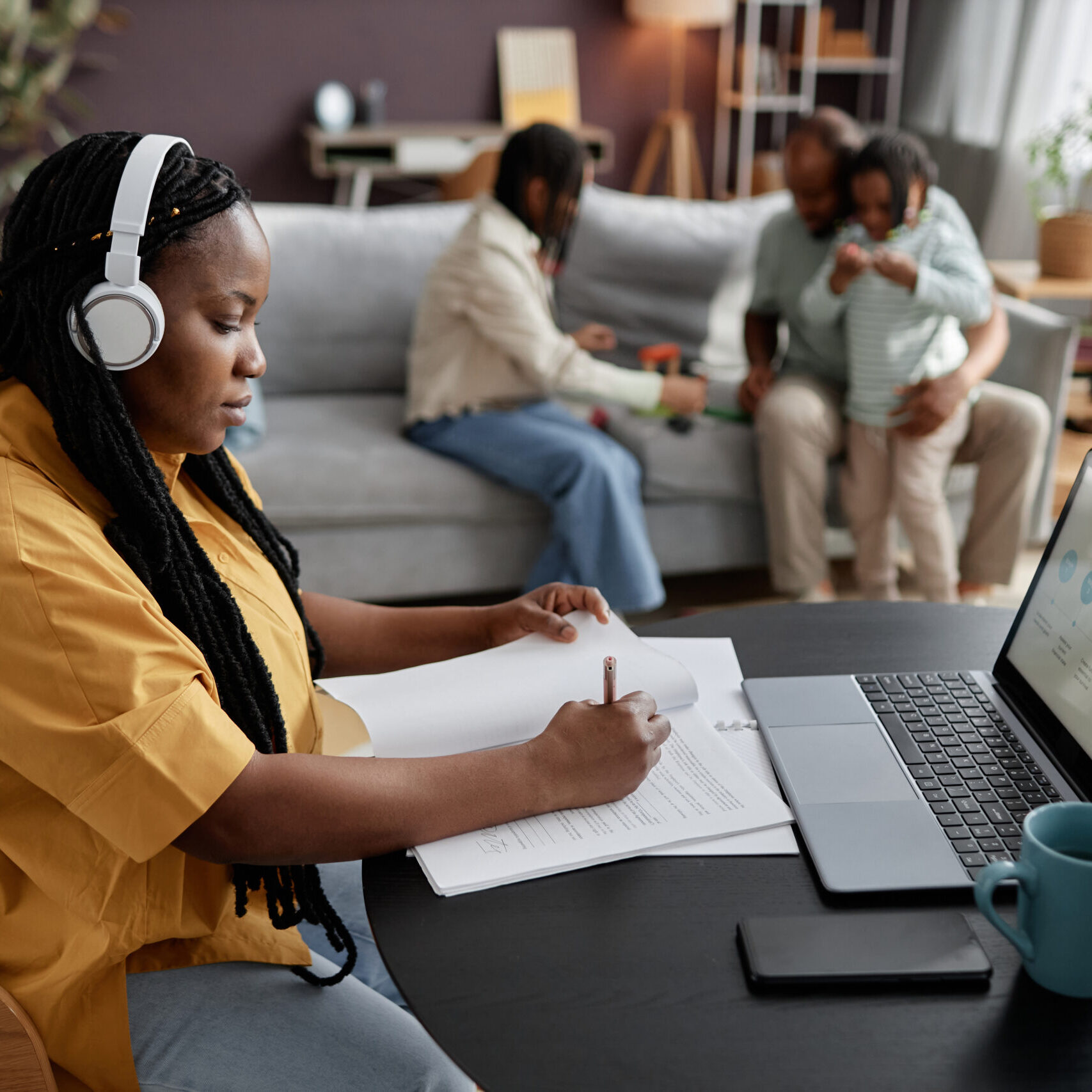 LRCE-Virtual-Program-Working-from-Home Woman wearing headphones working on laptop at table while family is relaxing on sofa in background. Taking notes and using technology in home workspace without distraction Working From Home While Family Relaxing in Background showing LRCE is a fully virtual program