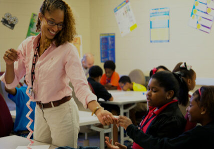 A smiling LRCE-certified teacher wearing a light pink shirt interacts with students in a classroom, handing a paper strip to a seated girl. Other students are engaged in activities at their desks in the background.