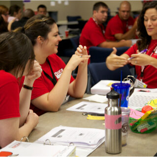 LRCE teachers getting certified A group of LRCE-certified educators in red shirts sit around a classroom table engaged in discussion during a training session. One woman gestures as she explains a concept while others listen and smile, surrounded by papers, binders, and teaching materials.
