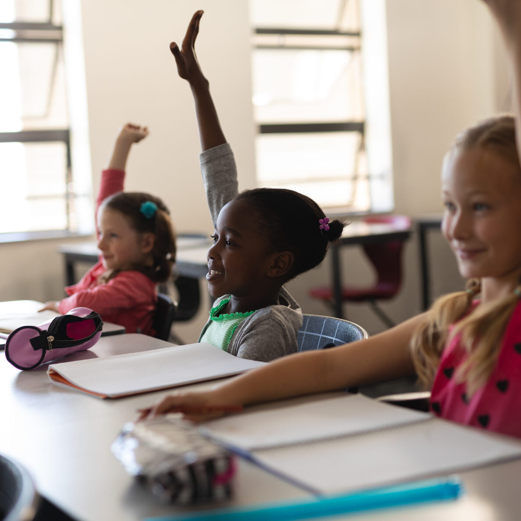 Three young students sit at their desks with notebooks open, smiling and eagerly raising their hands in a bright, engaging classroom—an environment inspired by LRCE-trained teachers.