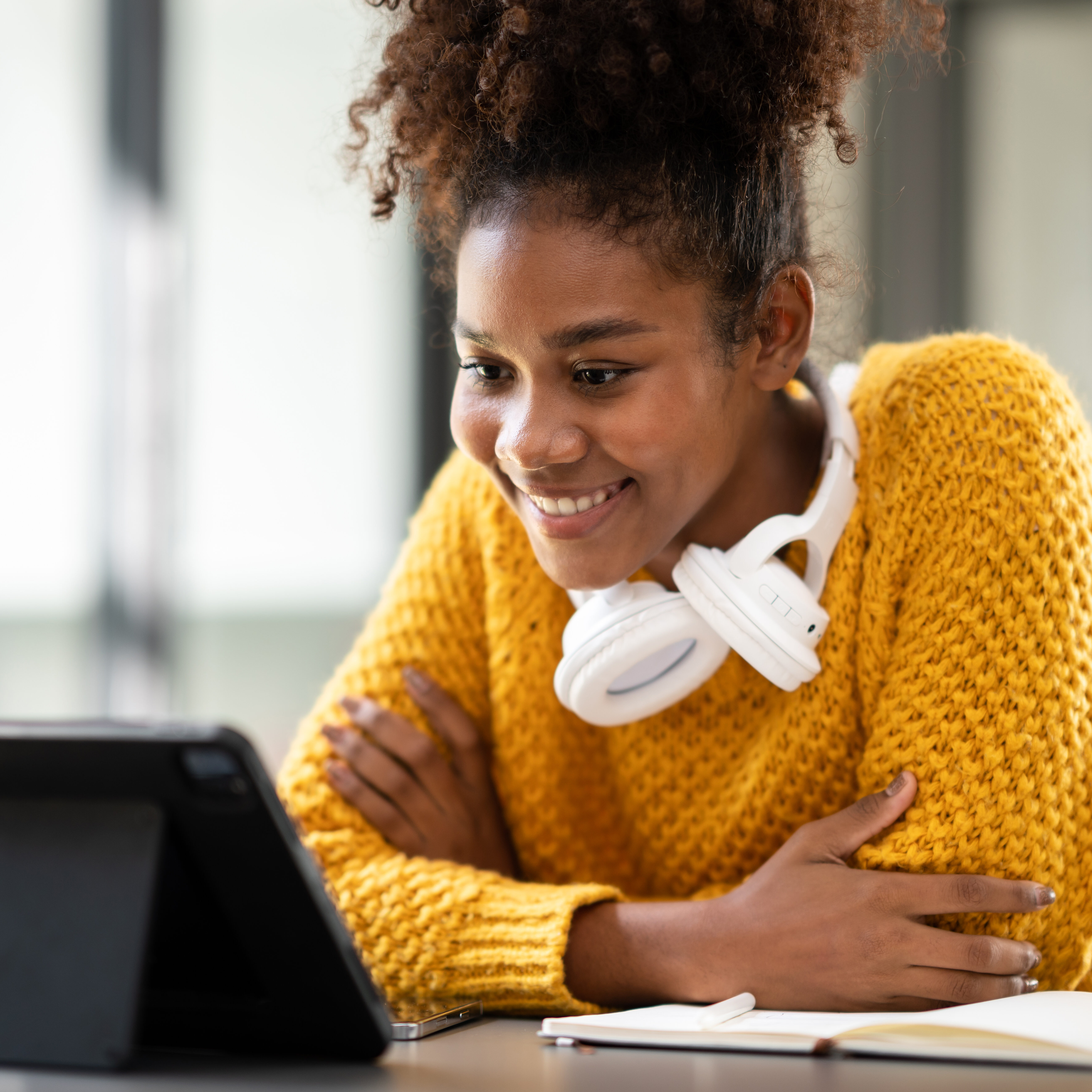 A smiling student wearing a yellow sweater and white headphones sits at a desk, engaging with content on a tablet. Her enthusiasm reflects the inclusive, student-centered impact of LRCE-trained educators and programs.
