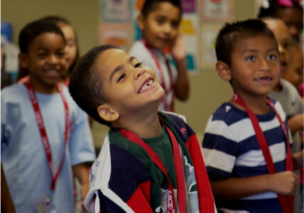 A group of young students smile and laugh together in a classroom, wearing red lanyards. Their joyful expressions reflect the positive and engaging environments created by LRCE-trained educators.