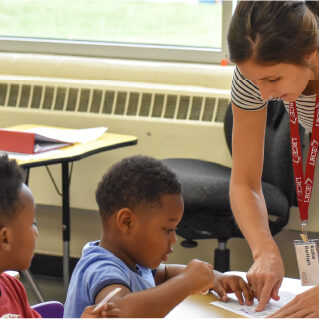 praxis-prep-support A teacher wearing an LRCE lanyard helps two young students with an assignment at their desk in a brightly lit classroom.